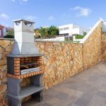 Barbecue en pierre maçonné sur une terrasse pavée moderne, avec un grand mur d'enceinte en pierres dorées et des habitations en arrière-plan sous un ciel bleu dégagé.