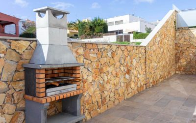 Barbecue en pierre maçonné sur une terrasse pavée moderne, avec un grand mur d'enceinte en pierres dorées et des habitations en arrière-plan sous un ciel bleu dégagé.