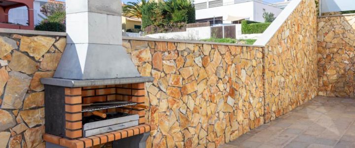 Barbecue en pierre maçonné sur une terrasse pavée moderne, avec un grand mur d'enceinte en pierres dorées et des habitations en arrière-plan sous un ciel bleu dégagé.
